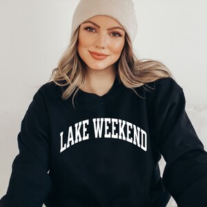 May include: A black sweatshirt with the text "LAKE WEEKEND" in white, arched lettering. The model is wearing a beige beanie.