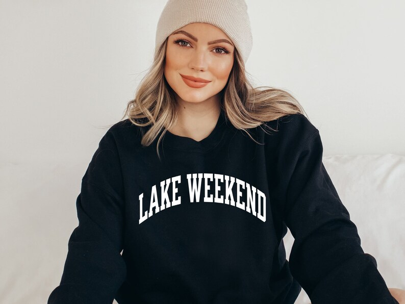 May include: A black sweatshirt with the text "LAKE WEEKEND" in white, arched lettering. The model is wearing a beige beanie.
