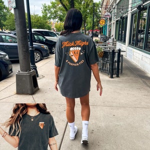 May include: A dark gray t-shirt with the text "Thick Thighs & Pumpkin Pies" and a cartoon pumpkin pie graphic. The shirt is being worn by a person with white socks and white sneakers.