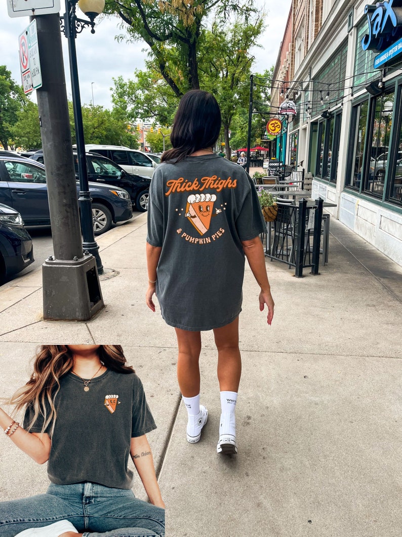 May include: A dark gray t-shirt with the text "Thick Thighs & Pumpkin Pies" and a cartoon pumpkin pie graphic. The shirt is being worn by a person with white socks and white sneakers.