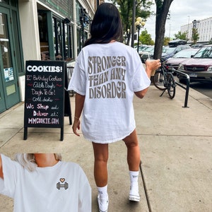 May include: White t-shirt with the text "STRONGER THAN ANY DISORDER" printed in brown on the back. A second white t-shirt with a small pink heart and a dumbbell graphic is shown in the foreground.