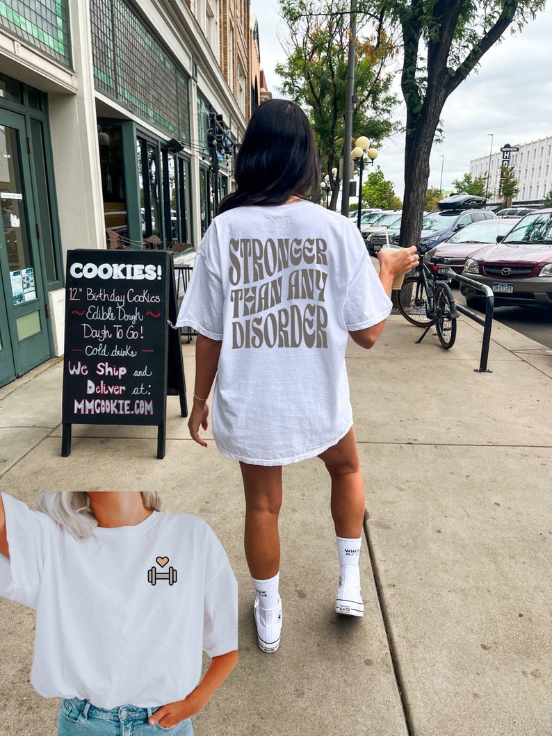 May include: White t-shirt with the text "STRONGER THAN ANY DISORDER" printed in brown on the back. A second white t-shirt with a small pink heart and a dumbbell graphic is shown in the foreground.
