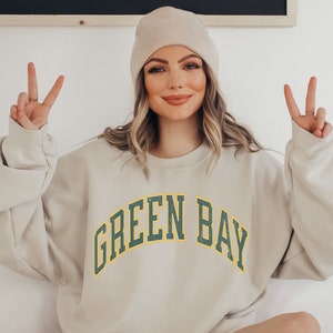 May include: A light beige sweatshirt with the text "GREEN BAY" in green and yellow letters. The sweatshirt has a relaxed fit and long sleeves.