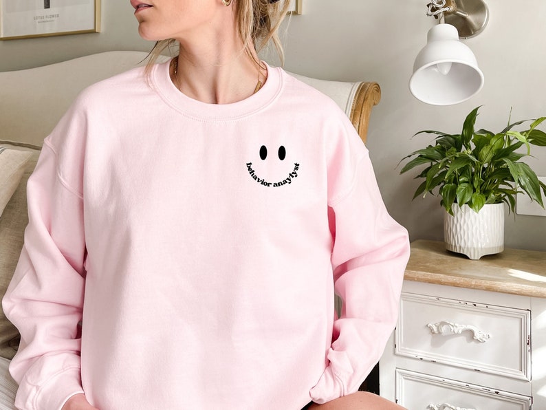 May include: Pink sweatshirt with a black smiley face and the text "behavior analyst" written inside the smile.