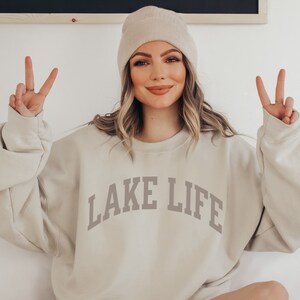 以下が含まれることがあります： ベージュのトレーナーに「LAKE LIFE」というグレーの文字が書かれたトレーナーを着た女性。ベージュのビーニーをかぶり、両手をピースサインにして掲げている。