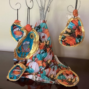 May include: Four handmade ornaments shaped like oyster shells with a fall theme. The ornaments are decorated with fabric featuring gnomes, pumpkins, and fall foliage. The ornaments are gold and blue with a white background.