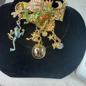 Puede incluir: Un broche detallado de rana dorada con una corona, posada en una hoja de nenúfar. El broche presenta dijes colgantes, que incluyen una pequeña rana, un orbe transparente y peces con gemas azules. La rana está pintada con esmalte verde, naranja y amarillo.