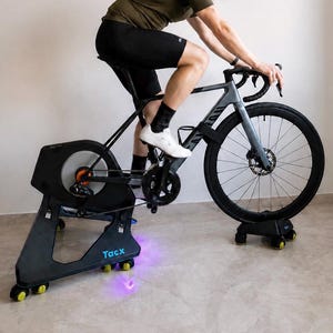 Op de afbeelding: Een grijze racefiets gemonteerd op een zwarte Tacx fietstrainer. De trainer heeft een groot cirkelvormig vliegwiel met een oranje midden en een basis met kleine gele wieltjes. De fiets is voorzien van zwarte banden en een schijfrem.