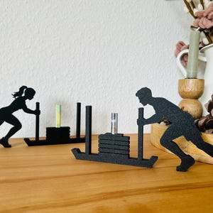 Könnte beinhalten: Zwei schwarze, texturierte Silhouettenfiguren von Athleten, die Gewichtsschlitten schieben. Jede Figur ist hinter einem Schlittenrahmen positioniert, mit einem zentralen vertikalen Pfosten, der gestapelte, rechteckige, schwarze Gewichtsplatten hält. Die Figuren befinden sich in einer nach vorne geneigten, athletischen Haltung, die eine Schiebebewegung nachahmt.
