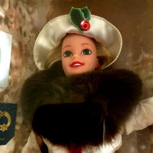 Puede incluir: Una muñeca vintage con un sombrero blanco con un adorno de baya de acebo, un cuello de piel oscura y un vestido blanco con botones rojos. La muñeca tiene cabello rubio, ojos verdes y lápiz labial rojo. La muñeca está en una caja.