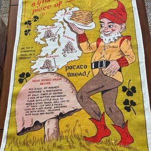 May include: A yellow linen tea towel featuring a map of Ireland, a leprechaun holding a plate of potato bread, and a printed recipe. The text reads: It's a Glorious piece of Ireland, Potato Bread, and an Irish Potato Bread recipe with measurements in ounces.
