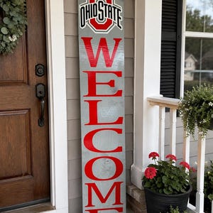 May include: A tall, rectangular wooden porch sign with a gray stained finish. The sign features the Ohio State University logo at the top in red, white, and black, followed by the word WELCOME written vertically in large, bold red capital letters.