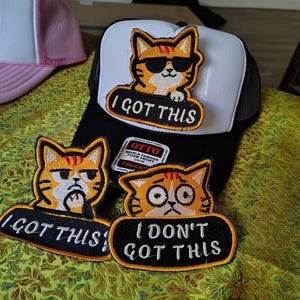 May include: A black and white trucker hat featuring an embroidered patch of an orange cat wearing sunglasses with the text I GOT THIS. Two additional patches are included: one showing a cat with the text I GOT THIS and one with I DON'T GOT THIS.