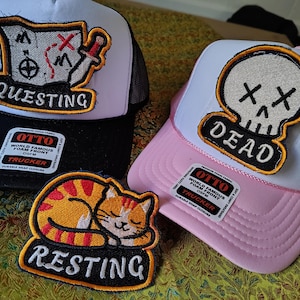 May include: Two trucker hats and one embroidered patch. One black and white hat features a map patch with the text QUESTING. One pink and white hat features a skull patch with the text DEAD. The patch shows a sleeping orange tabby cat with the text RESTING.