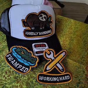 May include: A black and white trucker hat with a sloth patch reading HARDLY WORKING, accompanied by two separate patches: one shaped like an alligator reading SWAMPED and one with a wrench and pencil reading WORKING HARD.