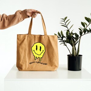 May include: A brown tote bag with a yellow smiley face dripping with black outlines. The bag has the text "Have a good trip." printed on the front.