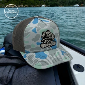 May include: A camouflage trucker hat with a blue and green pattern. The hat has a black and white fish graphic with the text "Only Fins" on the front.