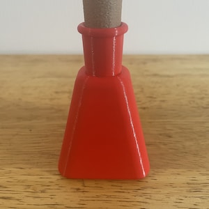 May include: A small, bright red glass bottle with a cork stopper. The bottle has a triangular shape and a narrow neck.