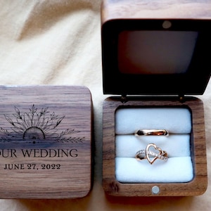 May include: A wooden ring box with a sunflower design and the text "OUR WEDDING JUNE 27, 2022". The box is open and contains two gold rings, one with a heart design.