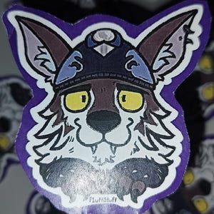 May include: A die-cut sticker featuring a stylized illustration of a wolf-like creature wearing a dark blue helmet with silver accents. The creature has large yellow eyes, white fur, and brown markings. The text FluffStuff is printed at the bottom.