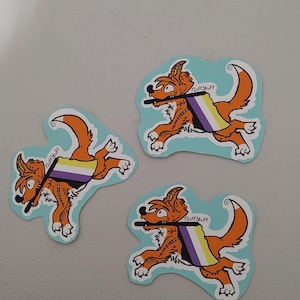 May include: Three die-cut stickers featuring an orange cartoon fox running while holding a non-binary pride flag in its mouth. The fox has white fur accents and the text FluffStuff is printed in black near the flag pole on each sticker.