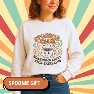 Puede incluir: Sudadera crema con el texto "Spoonie Club" en naranja y verde, con un gráfico de taza de café sonriente. Debajo del gráfico, el texto dice "Running on Empty Still Sparkling". Una pancarta naranja en la parte inferior dice "Spoonie Gift".