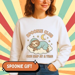 May include: White sweatshirt with a graphic of a sleeping sloth on a cloud, wearing a blue polka-dot onesie and nightcap. The text "Spoonie Club, One Nap at a Time" is above the sloth. An orange banner at the bottom says "Spoonie Gift".