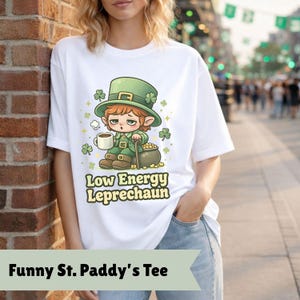 May include: White t-shirt with a cartoon leprechaun design. The leprechaun is wearing a green hat and holding a mug. Text on the shirt reads "Low Energy Leprechaun" and "Funny St. Paddy's Tee".