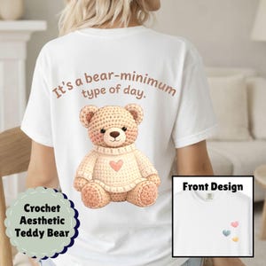 May include: White t-shirt featuring a crochet teddy bear design with a pink heart on the front. The back of the shirt has the text "It's a bear-minimum type of day." A small graphic of hearts is on the front. The shirt is part of the "Crochet Aesthetic Teddy Bear" collection.