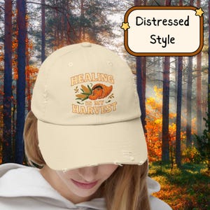 Könnte beinhalten: Cremefarbene Baseballkappe im Used-Look mit dem Text "HEALING IS MY HARVEST" in Orange und Braun. Das Design beinhaltet ein Füllhorn und Mais. Die Kappe hat einen gebogenen Schirm und ein abgenutztes Aussehen.