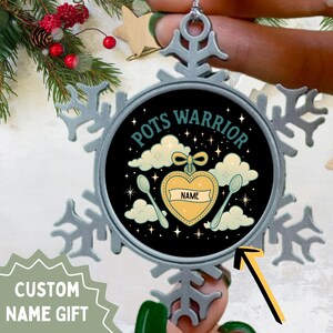 May include: A silver snowflake-shaped Christmas ornament with a circular centre. The centre has the words "Pots Warrior" and a design with a heart, spoons, and clouds. The word "NAME" is on the heart. The text "CUSTOM NAME GIFT" is also visible.