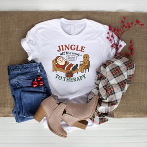 Peut inclure: T-shirt blanc avec un motif de Noël. Le motif représente le Père Noël sur un divan de thérapeute avec un bonhomme en pain d'épices. Le texte dit "Jingle all the way... to therapy". Le t-shirt est associé à un jean bleu, des bottines beiges et une écharpe à carreaux.