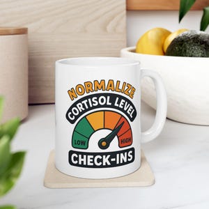 Può includere: Tazza in ceramica bianca con la scritta "NORMALIZE CORTISOL LEVEL CHECK-INS" in un design a indicatore. L'indicatore mostra livelli bassi, medi e alti. La tazza è su un sottobicchiere beige.