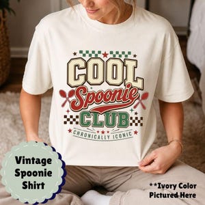 May include: Cream-coloured t-shirt with a vintage design. The shirt features the text "COOL Spoonie CLUB" in a retro font, with crossed spoons and stars. The phrase "Chronically Iconic" is also included.