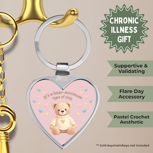 Puede incluir: Un llavero en forma de corazón con un fondo rosa que presenta un oso de peluche de ganchillo y el texto "It's a bear-minimum type of day." El llavero es un regalo para enfermedades crónicas.