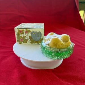 Może przedstawiać: Ozdobne pudełko z motywami kwiatowymi i ptasimi, obok zielonej szklanej miski wypełnionej ż&oacute;łtymi przedmiotami, prawdopodobnie cukierkami lub mydłami. Przedmioty są prezentowane na białym, okrągłym cokole na czerwonym tle.