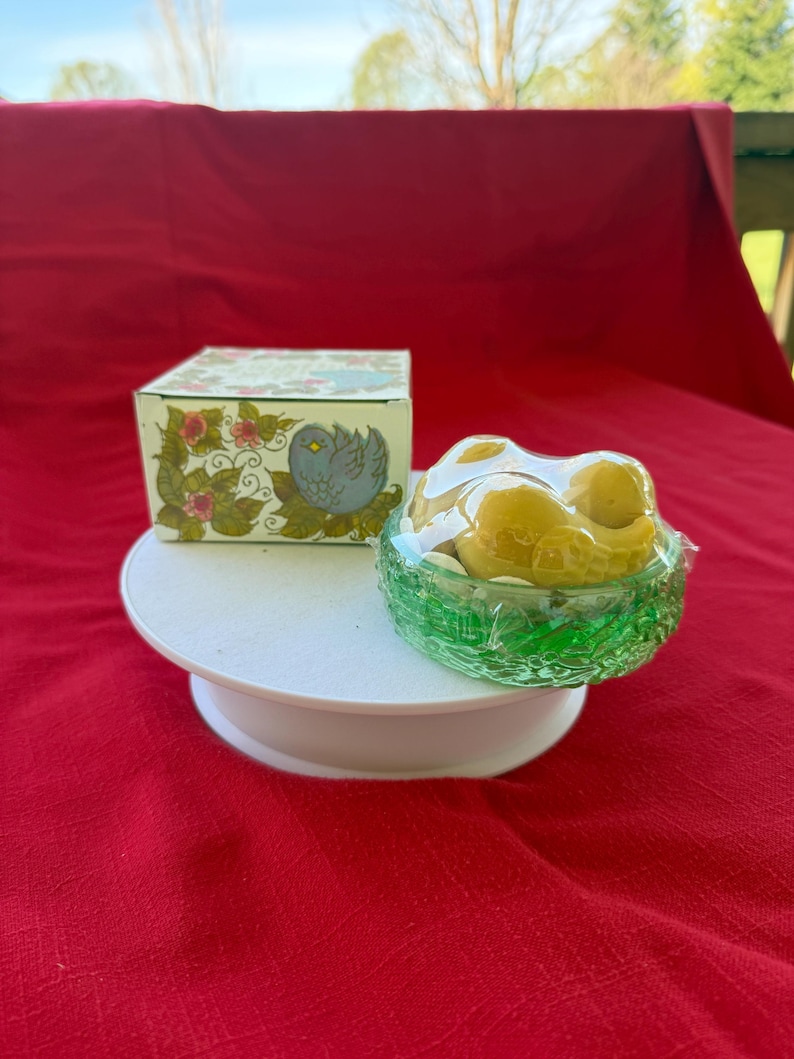 Może przedstawiać: Ozdobne pudełko z motywami kwiatowymi i ptasimi, obok zielonej szklanej miski wypełnionej ż&oacute;łtymi przedmiotami, prawdopodobnie cukierkami lub mydłami. Przedmioty są prezentowane na białym, okrągłym cokole na czerwonym tle.