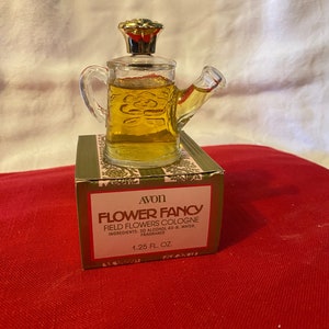 May include: A vintage Avon Flower Fancy cologne bottle shaped like a watering can, filled with amber liquid. The bottle sits on a green and red box with the product name and ingredients. The bottle is 1.25 fl oz.