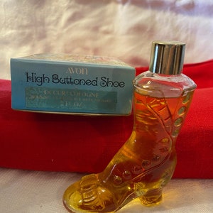 May include: A clear glass bottle shaped like a high-heeled shoe filled with yellow liquid. The bottle has a gold cap and is labeled "Avon High Buttoned Shoe Occur! Cologne 2 fl oz".