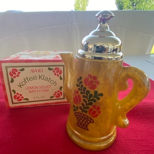 May include: A vintage Avon "Koffee Klatch" bath foam bottle in the shape of a yellow teapot with a silver lid. The teapot is decorated with red flowers and green leaves. The box next to it reads "Lemon Velvet Bath Foam" and "5 fl oz."