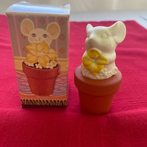 May include: A white mouse figurine with a yellow flower, sitting in a small orange pot. A matching box with a mouse illustration and floral design is next to the figurine. The background is a red surface.