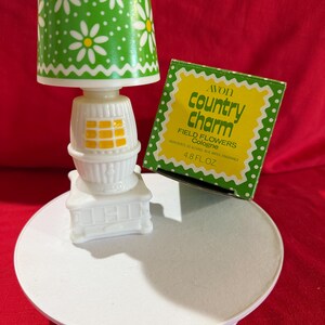 May include: A vintage white and green decorative lamp with a daisy pattern on the shade, and a small box of Avon Country Charm Field Flowers Cologne. The lamp is on a white pedestal.