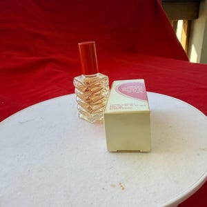 May include: A vintage perfume bottle with a red cap and a clear, textured glass body. The bottle is next to a small, off-white box with pink text. The items are on a white surface against a red backdrop.