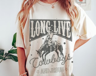 Könnte beinhalten: Cremefarbenes T-Shirt mit einem verblichenen Grafikdesign. Es zeigt einen Cowboy auf einem Pferd, mit dem Text "LONG LIVE Cowboys AND JESUS". Das Design ist in Grau- und Schwarztönen gehalten, mit Sternen.