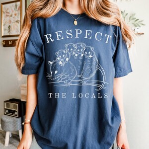 Puede incluir: Camiseta azul marino con la frase blanca "RESPECT THE LOCALS". La camiseta presenta un gr&aacute;fico de zarig&uuml;eyas. Una prenda de estilo casual.