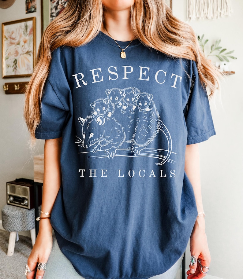 Puede incluir: Camiseta azul marino con la frase blanca "RESPECT THE LOCALS". La camiseta presenta un gr&aacute;fico de zarig&uuml;eyas. Una prenda de estilo casual.
