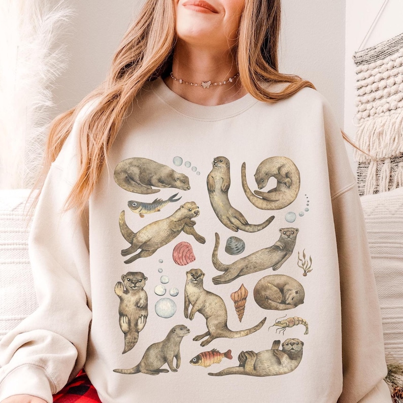 Peut inclure: Sweat-shirt beige avec une illustration &agrave; l'aquarelle de loutres dans diverses poses, avec des bulles, des coquillages et des poissons. Les loutres sont repr&eacute;sent&eacute;es dans des tons bruns et gris, cr&eacute;ant un motif sur le th&egrave;me de la nature.