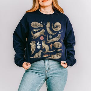Peut inclure: Sweat-shirt bleu marine orn&eacute; d'un motif d'otaries dans diverses poses, avec des coquillages et des poissons. Les loutres sont repr&eacute;sent&eacute;es dans des tons bruns et gris, cr&eacute;ant un design fantaisiste. Le sweat-shirt est de style d&eacute;contract&eacute;.