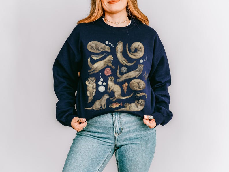 Peut inclure: Sweat-shirt bleu marine orn&eacute; d'un motif d'otaries dans diverses poses, avec des coquillages et des poissons. Les loutres sont repr&eacute;sent&eacute;es dans des tons bruns et gris, cr&eacute;ant un design fantaisiste. Le sweat-shirt est de style d&eacute;contract&eacute;.