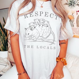 Puede incluir: Camiseta blanca con un gr&aacute;fico de una zarig&uuml;eya y sus cr&iacute;as, con las palabras "RESPECT THE LOCALS" en letras grises. La ilustraci&oacute;n de la zarig&uuml;eya es detallada y el texto est&aacute; en una fuente sencilla. La camiseta es de estilo casual.